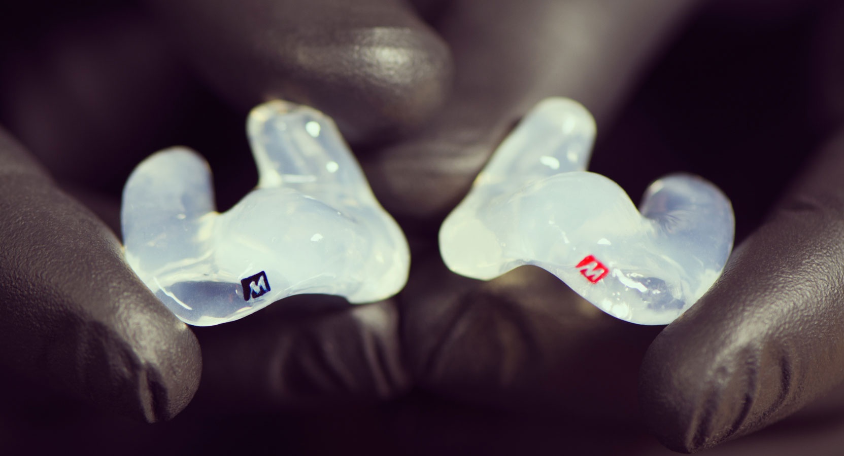 Hand wearing black nitrile gloves holding a pair of custom silicone eartips in clear color showing off the logos